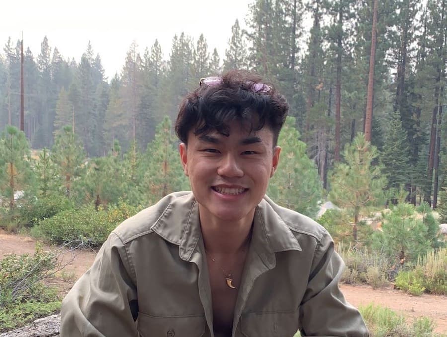 Man in a beige button down and gold necklace smiling in the woods on a foggy day. 