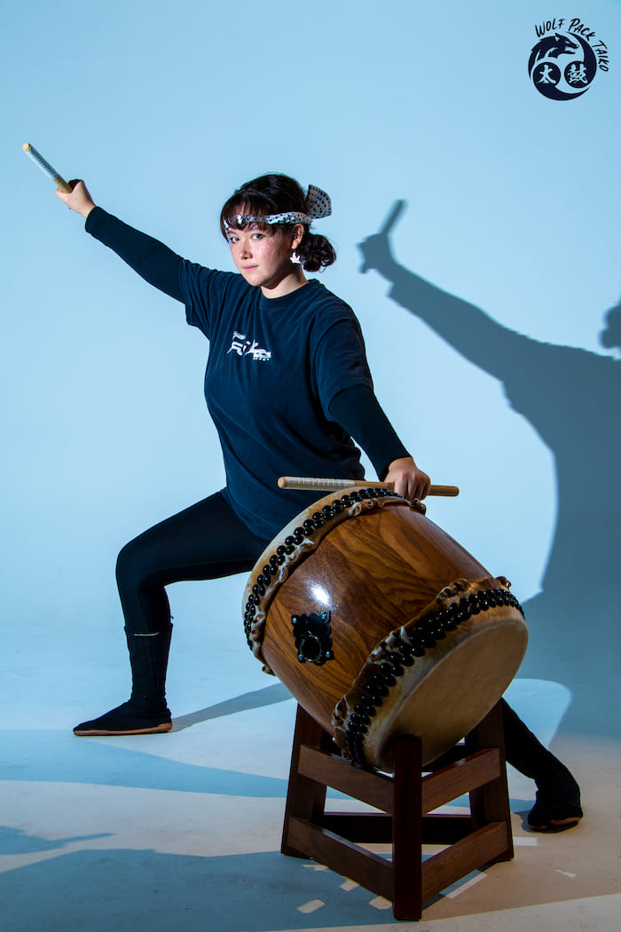 Girl with a bandana in front of light blue background wearing all black posing with two drumsticks in her hands and one arm above her head with the other reaching down to the brown drum in front of her on the floor.