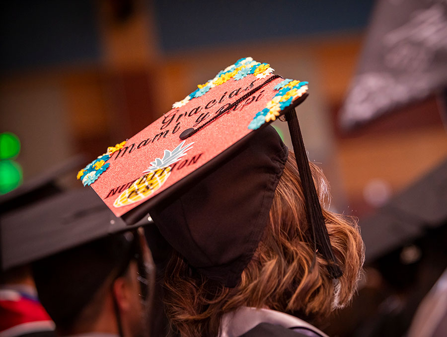 A student is pictured wearing a mortar board cap that reads "Gracias mami y papi" on the top of the cap and "Nutrition B.S." on the bottom of the cap with a pineapple illustration beneath it 