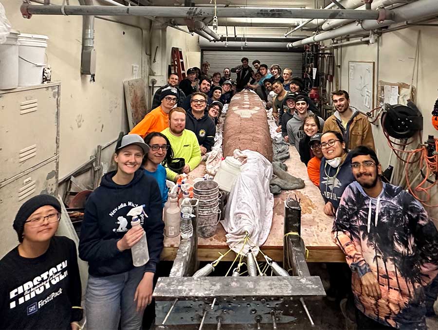 31 students in a garage setting standing around a mold of canoe, ready to pour concrete.