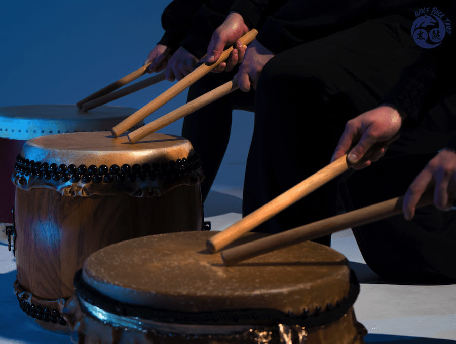 Close up of three people wearing all black and playing tall drums that reach just below their waists with light brown drumsticks in front of a blue background. 