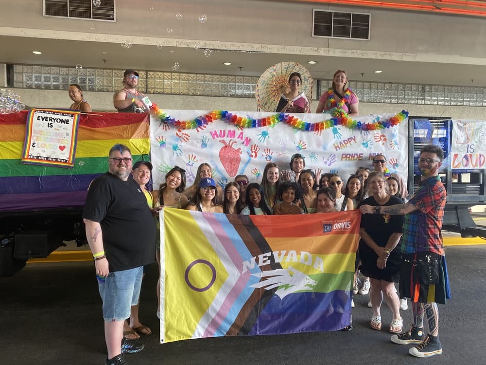 A group of 25 students, faculty and friends standing in front of a float decorated for pride, holding a pride flag