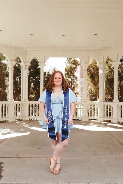 Addie Heithecker stands under a gazebo in a dress and graduation stoles