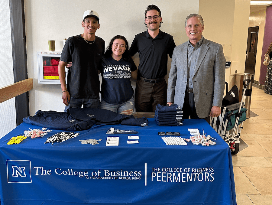 Three College of Business students posing with Dean Greg Mosier behind a decorated College of Business booth