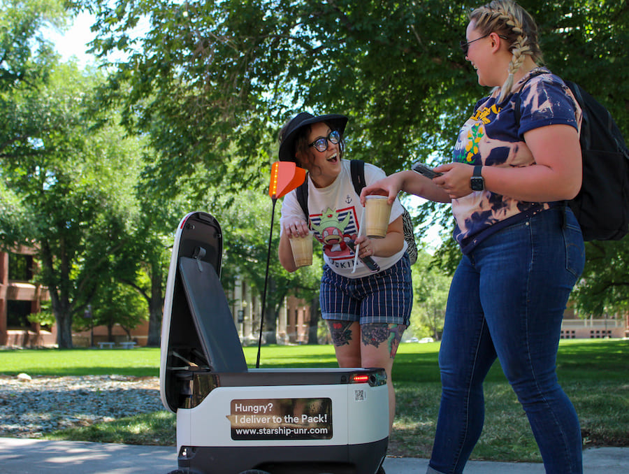 Two smiling students who are holding coffee that they just pulled out of a small, white delivery robot with the words "Hungry? I deliver to the pack! starship-unr.com"