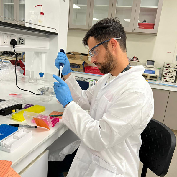 A man in a lab coat and safety glasses sits at a lab bench with a pipette in his hand.