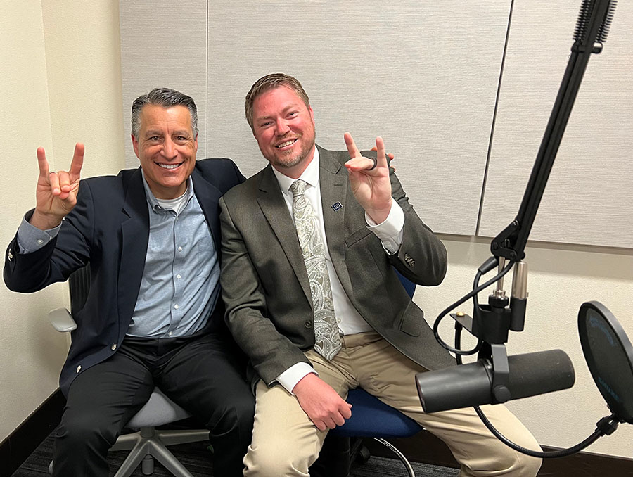 President Brian Sandoval sits next to Dr. Jake DeDecker in the podcasting room at the Reynold's School of Journalism. Both smile broadly with podcasting microphones in front of them, using their hands to make the Wolf Pack hand signal.