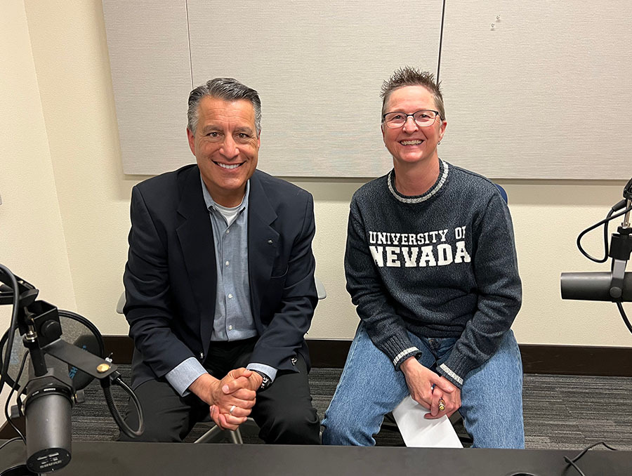 President Brian Sandoval sits next to Dr. Jill Heaton in the podcasting room at the Reynold's School of Journalism. Sandoval wears a suit and Heaton wears a Nevada Sweater and both smile broadly with podcasting microphones in front of them.