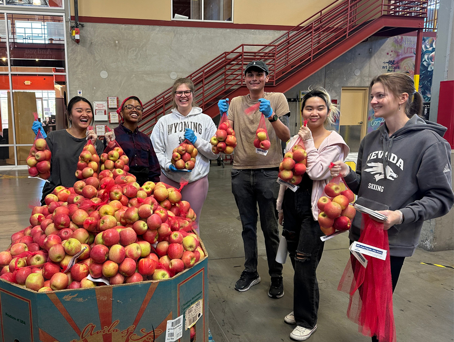 Beta Alpha Psi team holds bags of apples next to a produce container full of apples while volunteering at the Food Bank of Northern Nevada.