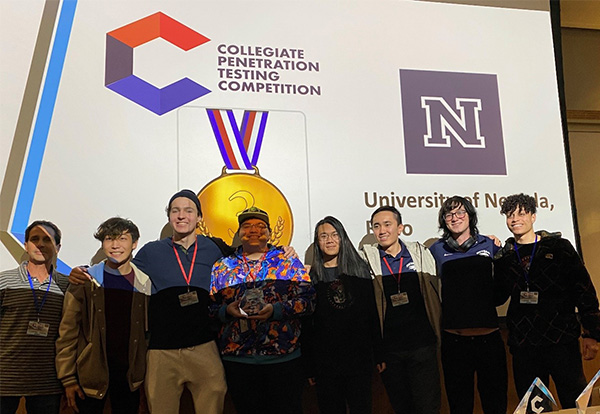 Eight students stand in front of a large projector image that reads College Penetration Testing Competition