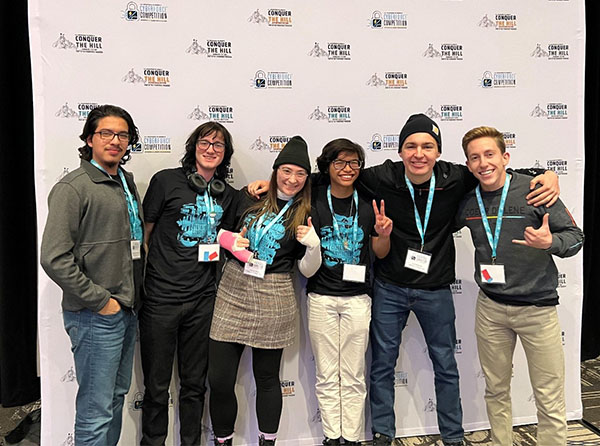 Six students stand in front of a photo backdrop that has the logo Conquer the Hill CyberForce competition repeated multiple times.