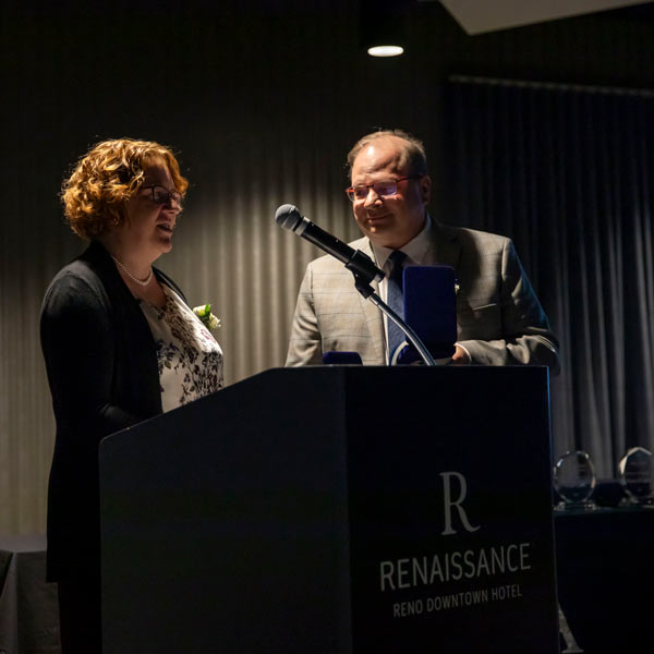 Leslie Sherrill speaks at a podium while Manolo Sherrill looks at her.