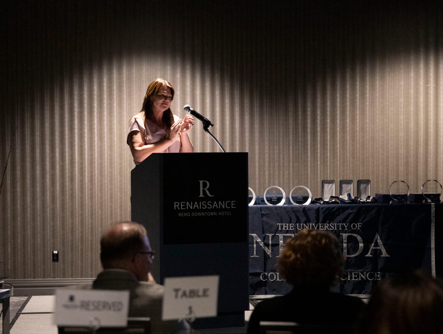 Louisa Hope-Weeks addresses an audience at a podium with her hands clasped. There are glass awards on a table behind her.