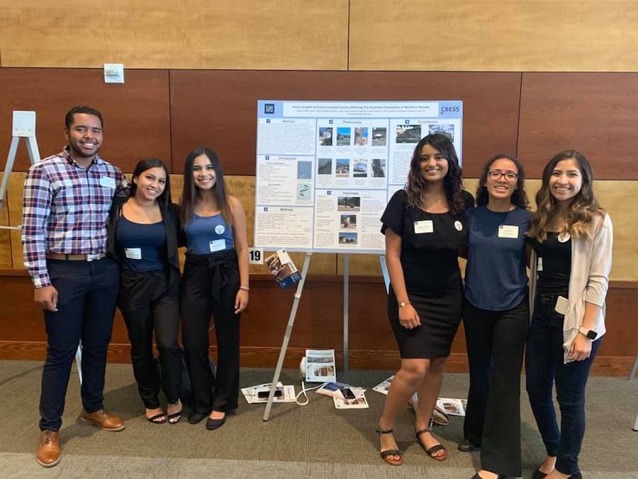 Six people stand in business attire, three on one side, and three on the other of a research poster, smiling.