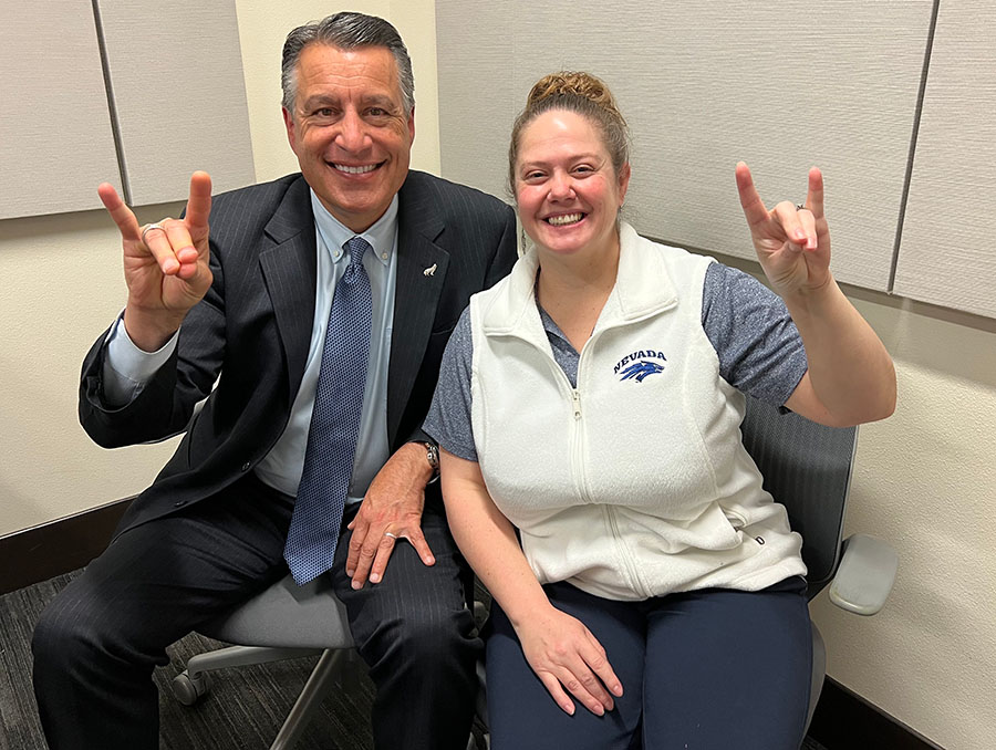 President Brian Sandoval sits next to Jessica Keefhaver in a podcasting studio. Both people hold their hands in the wolf sign.