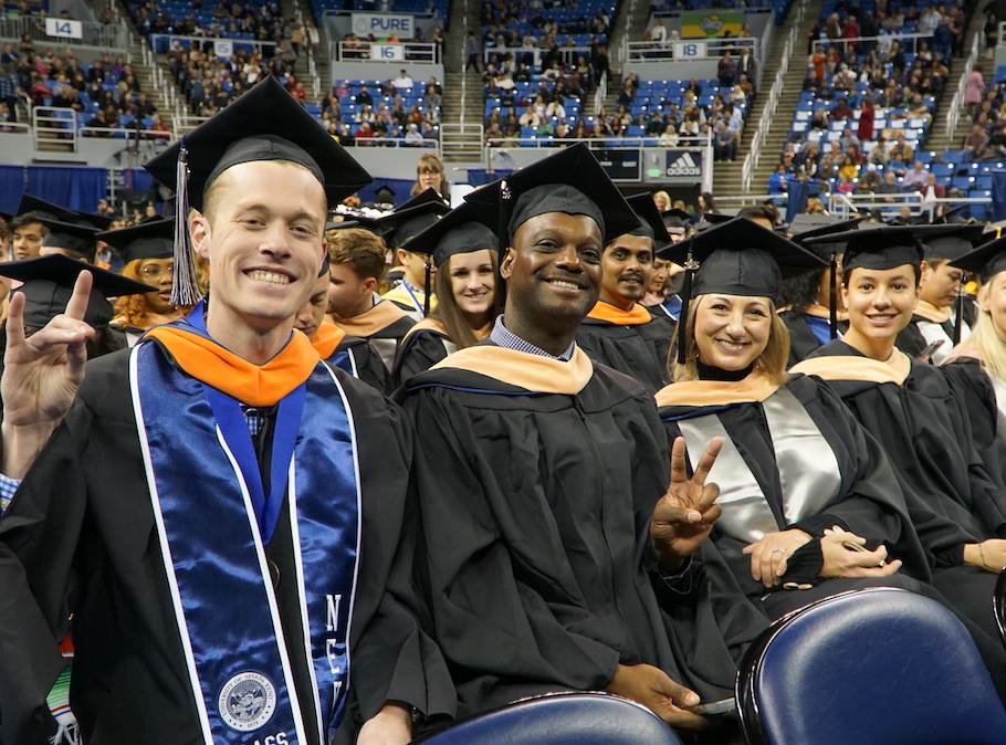 Four students smile at the camera in master's degree regalia on the floor of Lawlor during winter commencement. The one closest to the camera makes the Wolf Pack hand sign.