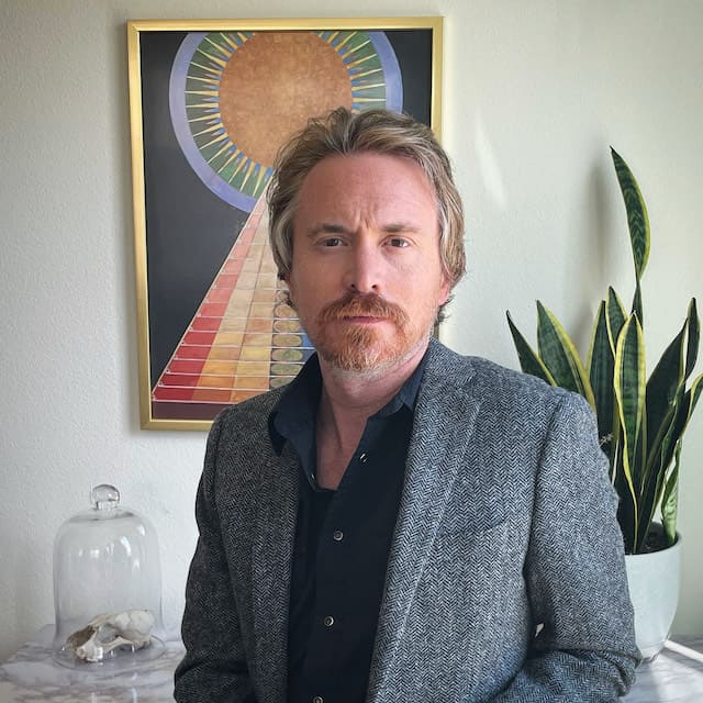 Man in front of painting and plant in a grey suit and black button-up, collared shirt.