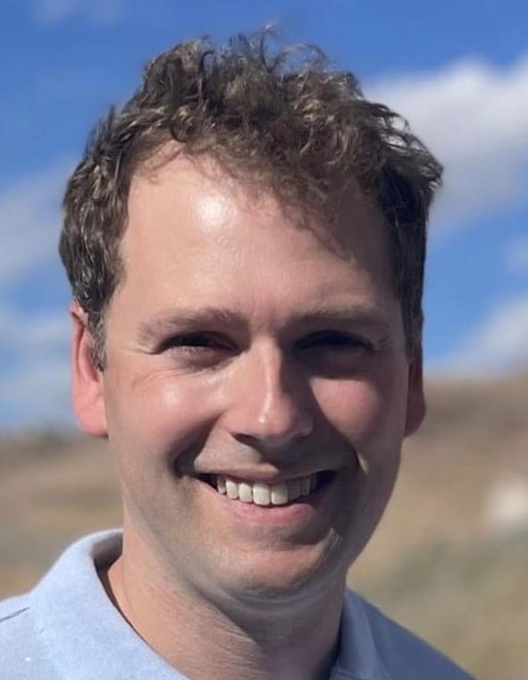 Man smiling against a blue sky background in a blue-collared shirt..