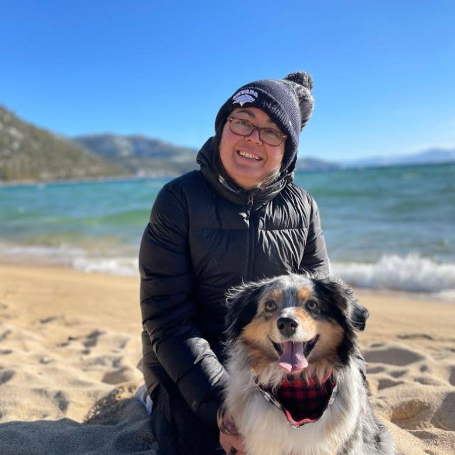 A woman wearing a coat and a blue UNR beanie sits on a beach at Lake Tahoe with a mini Australian Shepherd.