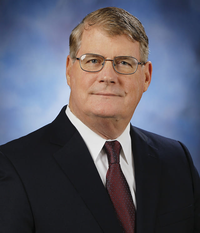 Man in front of a blue and white background in a black suit, white collared button-up and red tie.