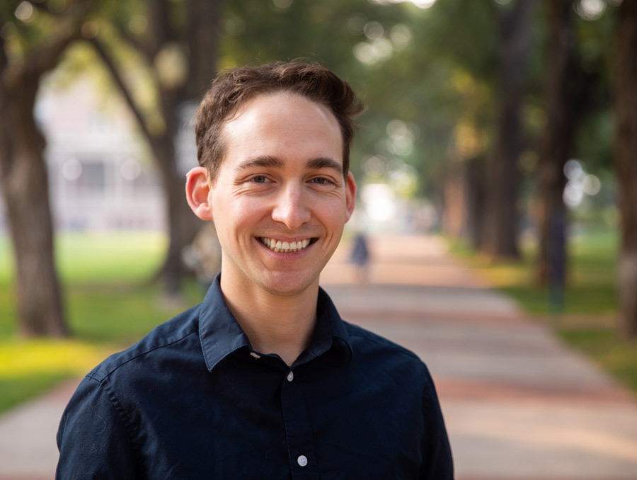 Thomas White, wearing a dark blue button-up collared shirt, smiles on the Quad.