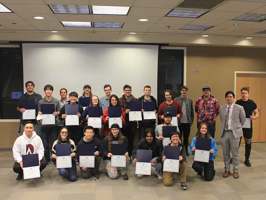 Twenty-three people standing inside a room in front of all wall-sized projector screen, lined up in three rows. Most are holding certificates facing toward the camera.