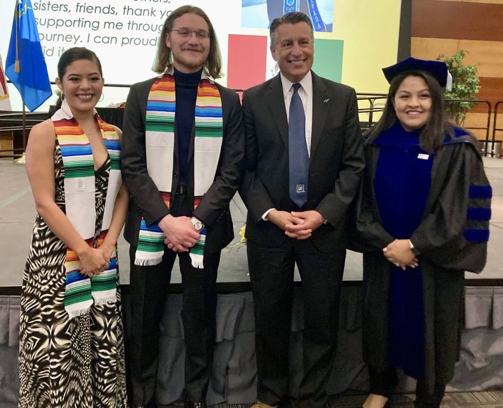 Two students, administrative faculty and University President smile in front of a stage at the Latinx Graduation Celebration with all four dressed in graduation gowns and caps. 