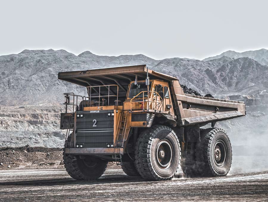 A large mining haul truck drives toward the left of the photo. There are large mountains in the background.
