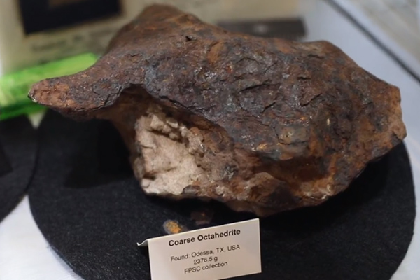 A brown rock with a lighter interior sits on a black felt pad. A label reads, "Coarse Octahedrite. Found: Odessa, TX, USA, 2376.5 g, FPSC collection"