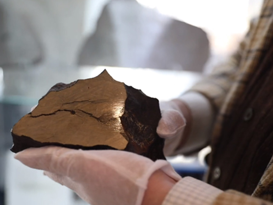 A person wearing gloves holds a rock in their hand. The rock is dark gray on the exterior, but the interior is cut and is a shiny bronze color.