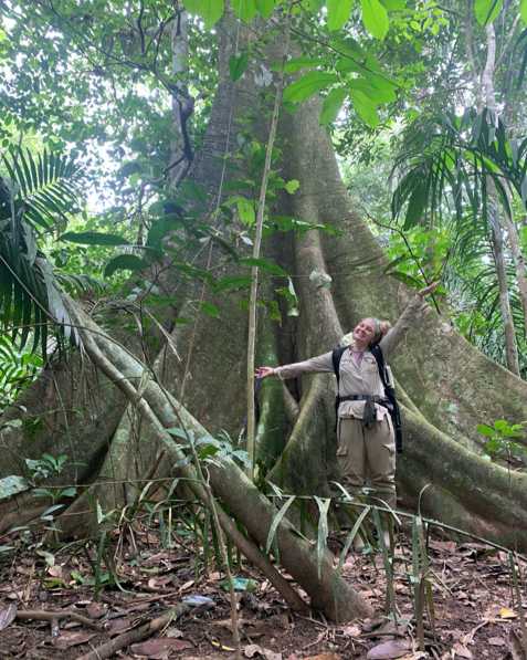 A girl wearing a backpack stands in front of a large tree in a tropical setting with her arms up, smiling.