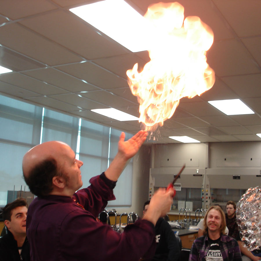 A man wearing a red collared shirt gives a demonstration in a classroom, holding a lighter in one hand and fire in his other hand.