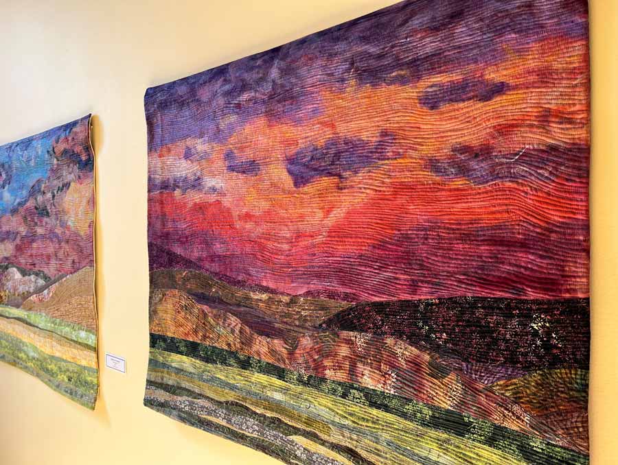 Two colorful quilts hang on a wall. They depict mountainous Nevada landscapes at sunset.