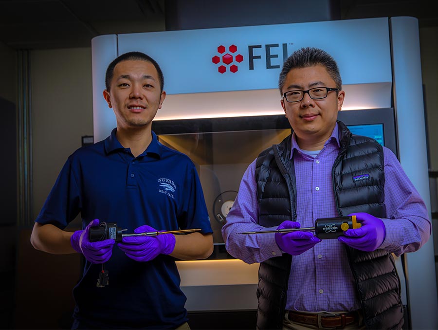 Dian Li and Yufeng Zheng standing in front of a electron microscope, holding equipment.