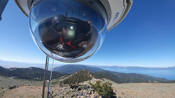 A temperature sensor with a glass dome is seen above a desert landscape with Lake Tahoe in the background. Researcher Scotty Strachan is reflected in the glass dome, as he is the one taking the picture.