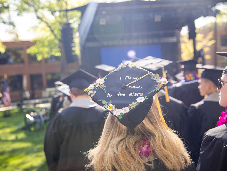 At the University graduation on the Quad, a student's cap is in focus, reading "I flew now, to the stars"