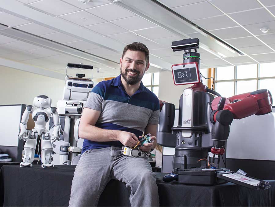 David Feil-Seifer sitting on a table next to robots.