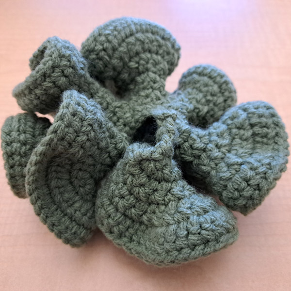 A crocheted structure that looks similar to a flower sits on a wooden desk. It is made with green yarn.