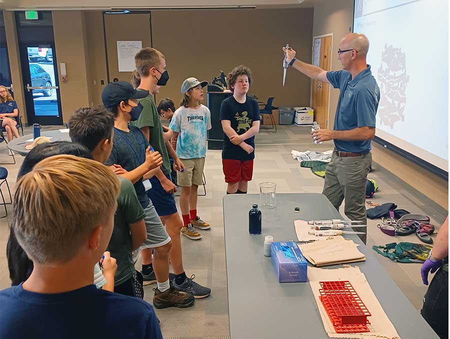 Eight middle school-aged youth in a classroom looking at a professor, who is holding lab equipment.