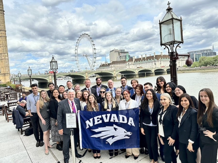 A group of 26 students and three professors pose with a Wolf Pack flag and the London Eye ferris wheel in the background. 
