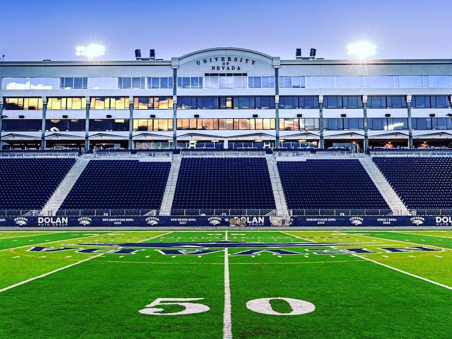Mackay stadium as seen from the 50 yard line of the football field just as evening falls and the stands are illuminated but empty. The words "University of Nevada" are inscribed on the building above the bleachers. 