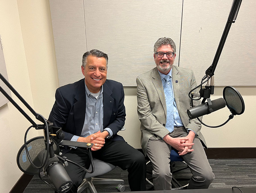 President Sandoval sits to the left of Peter Reed in a podcast recording room.
