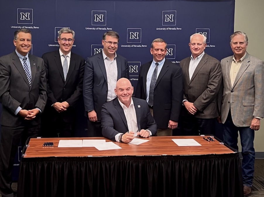 A group of men in business suits stand around a table as one man sits and signs multiple papers. All are smiling. 