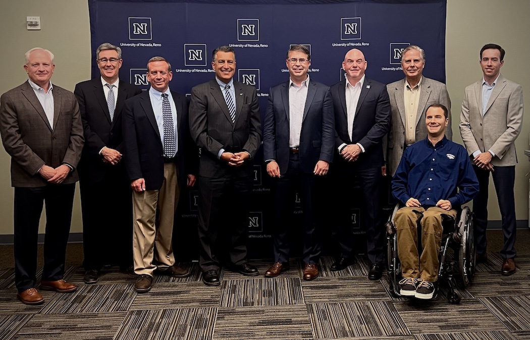 A group of individuals in business suits smile for the camera with a step and repeat backdrop with the University of Nevada, Reno's logo on it.