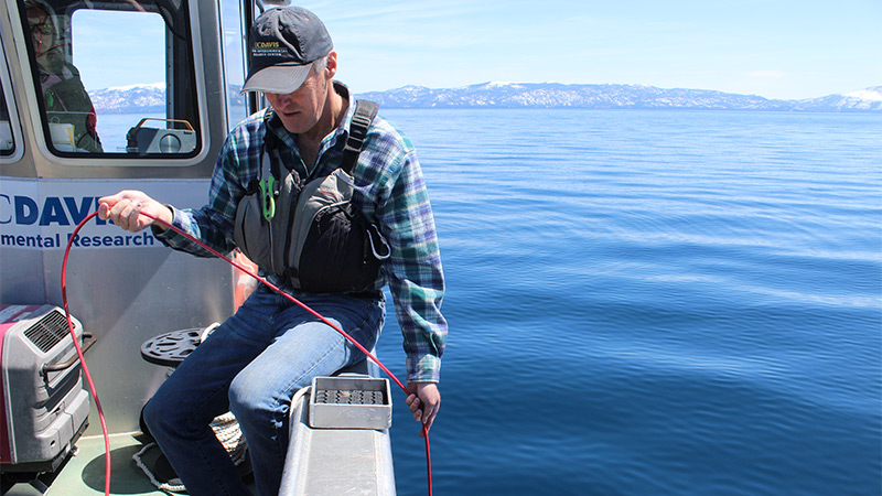 A man sits on the edge of a boat and lowers a device on a red rope into the water to measure the lake's clarity