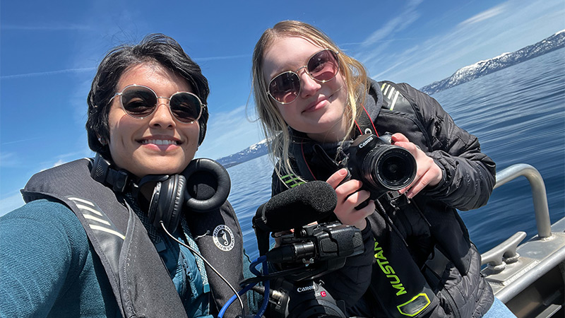 Two students on a boat wearing life vests and holding video and audio equipment