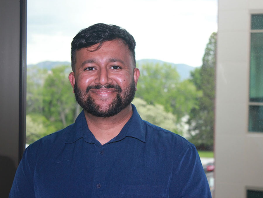 Portrait of Aditya Nair in a blue shirt standing outside, buildings and foliage in the background.