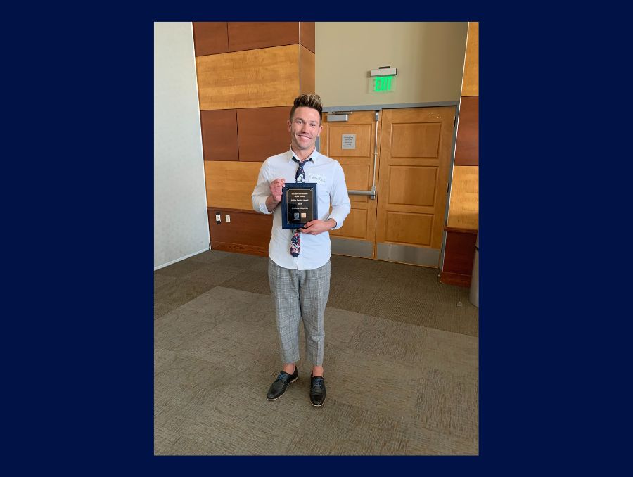 Matthew Kopicko stands holding his award in a dress shirt and pants with a floral tie. 