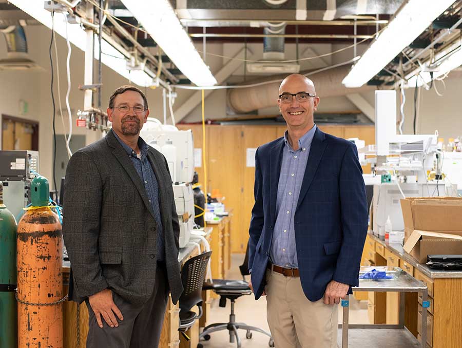 Two men in suits standing in a lab and looking at the camera.