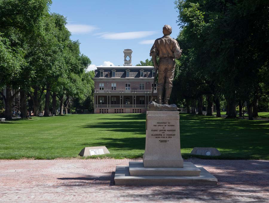 The back of the Mackay statue facing toward the Quad. The grass is green and the trees lining the Quad are fully leafed out. The sky is blue with some scattered clouds.